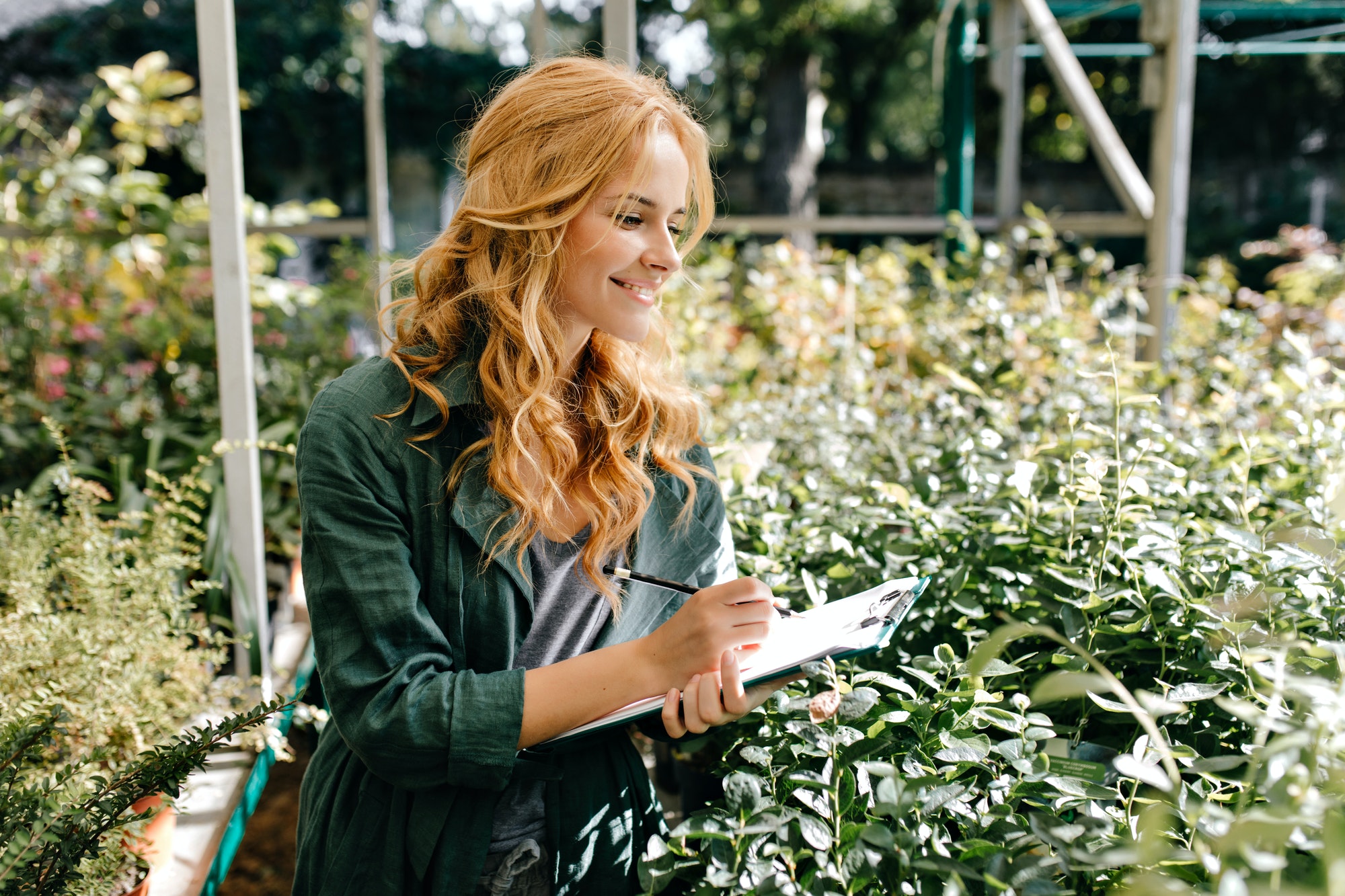 Garden filled with live plants. Young girl with beautiful blond hair and gentle smile, dressed in g